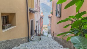 A narrow cobblestone street flanked by buildings with balconies, leading to a body of water and mountains in the distance. - Casa La Nuvola Varenna (Varenna)