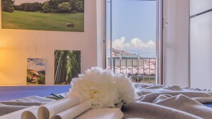 A bed with a book and a white flower, a window with a view of a building and trees, and artwork on the wall. - Casa La Nuvola Varenna (Varenna)
