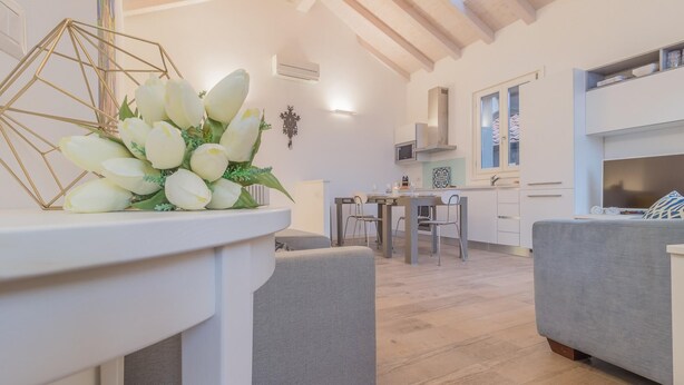 Un salon moderne avec un coin repas, comprenant une table blanche agrémentée d’un bouquet de fleurs blanches, des canapés gris et une cuisine en arrière-plan.