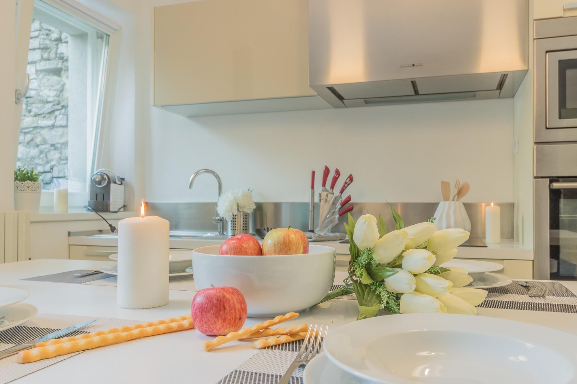 A modern kitchen with a white countertop, a white bowl with apples and tulips, a lit candle, and a window with a stone wall view.