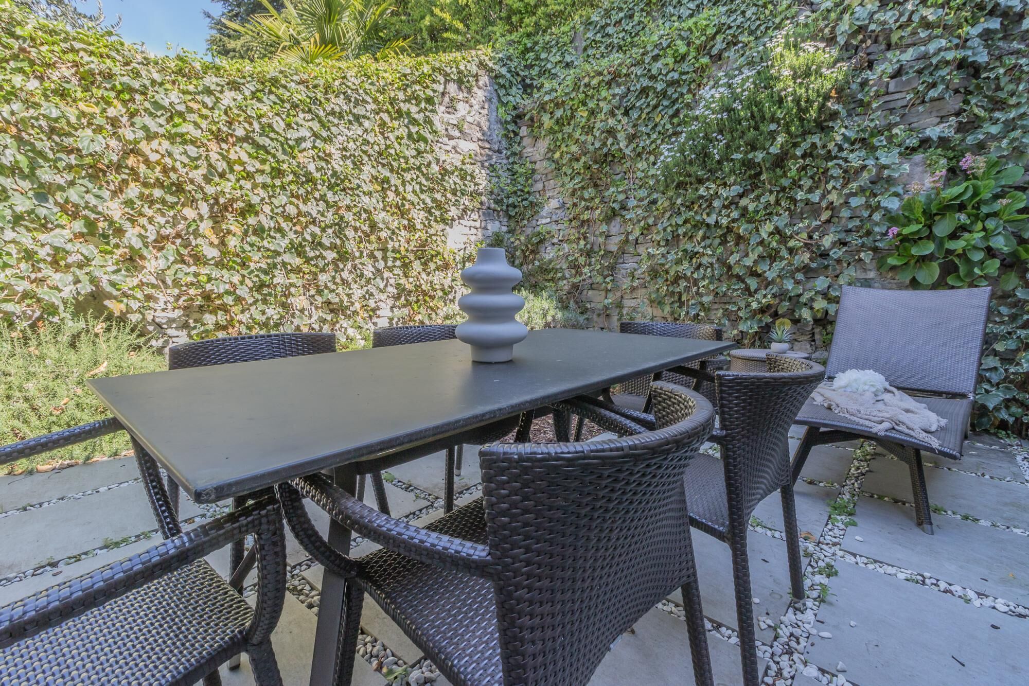 Un patio al aire libre con una mesa y sillas de mimbre, rodeado por un muro de piedra cubierto de hiedra.