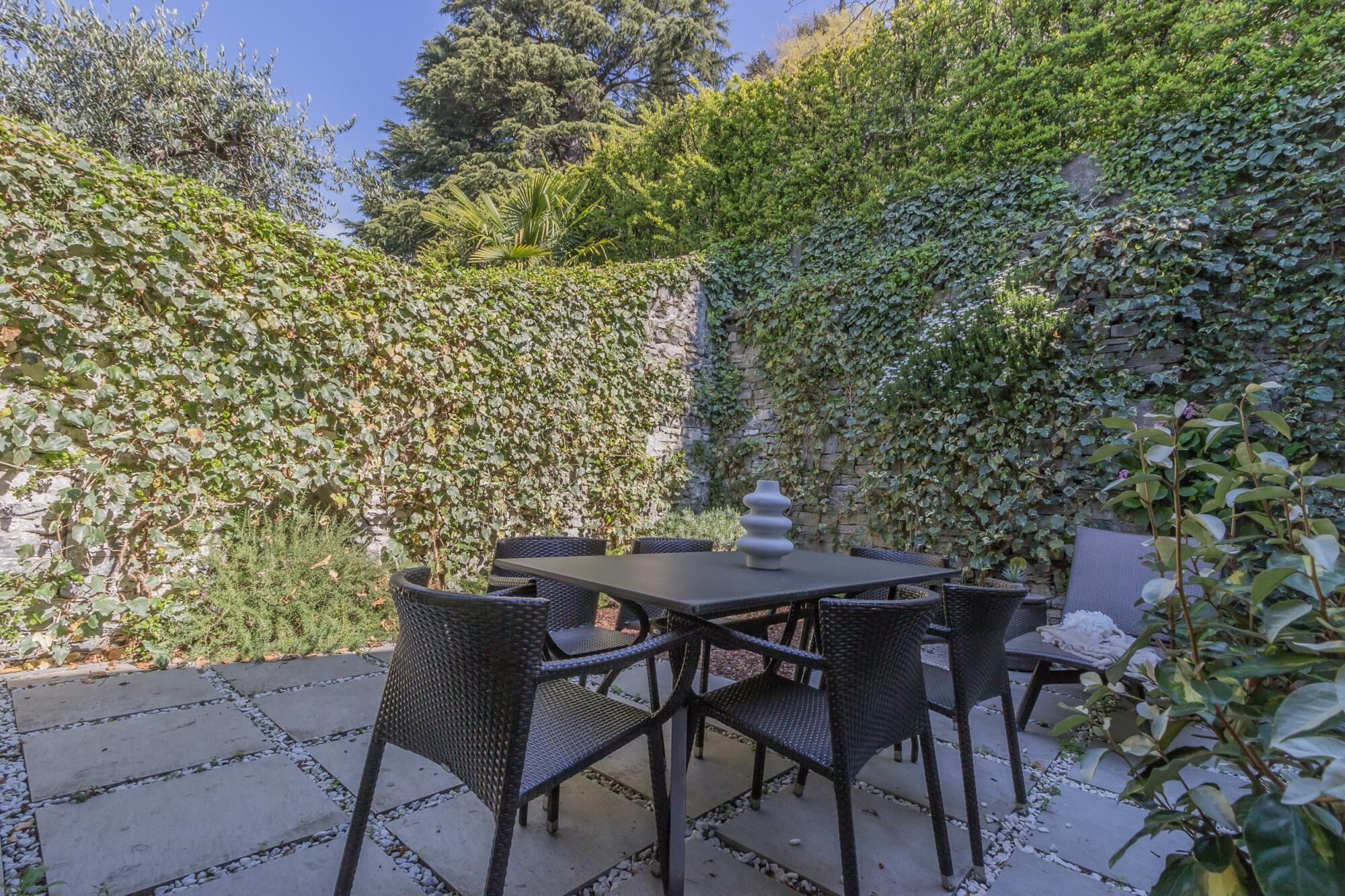 An outdoor patio with a table and chairs, surrounded by lush greenery and a stone wall.