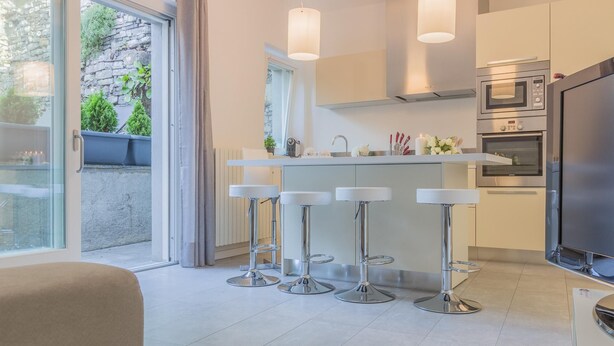Una cocina moderna con horno empotrado, microondas y un televisor grande. Hay cuatro taburetes de bar con asientos blancos y bases cromadas.