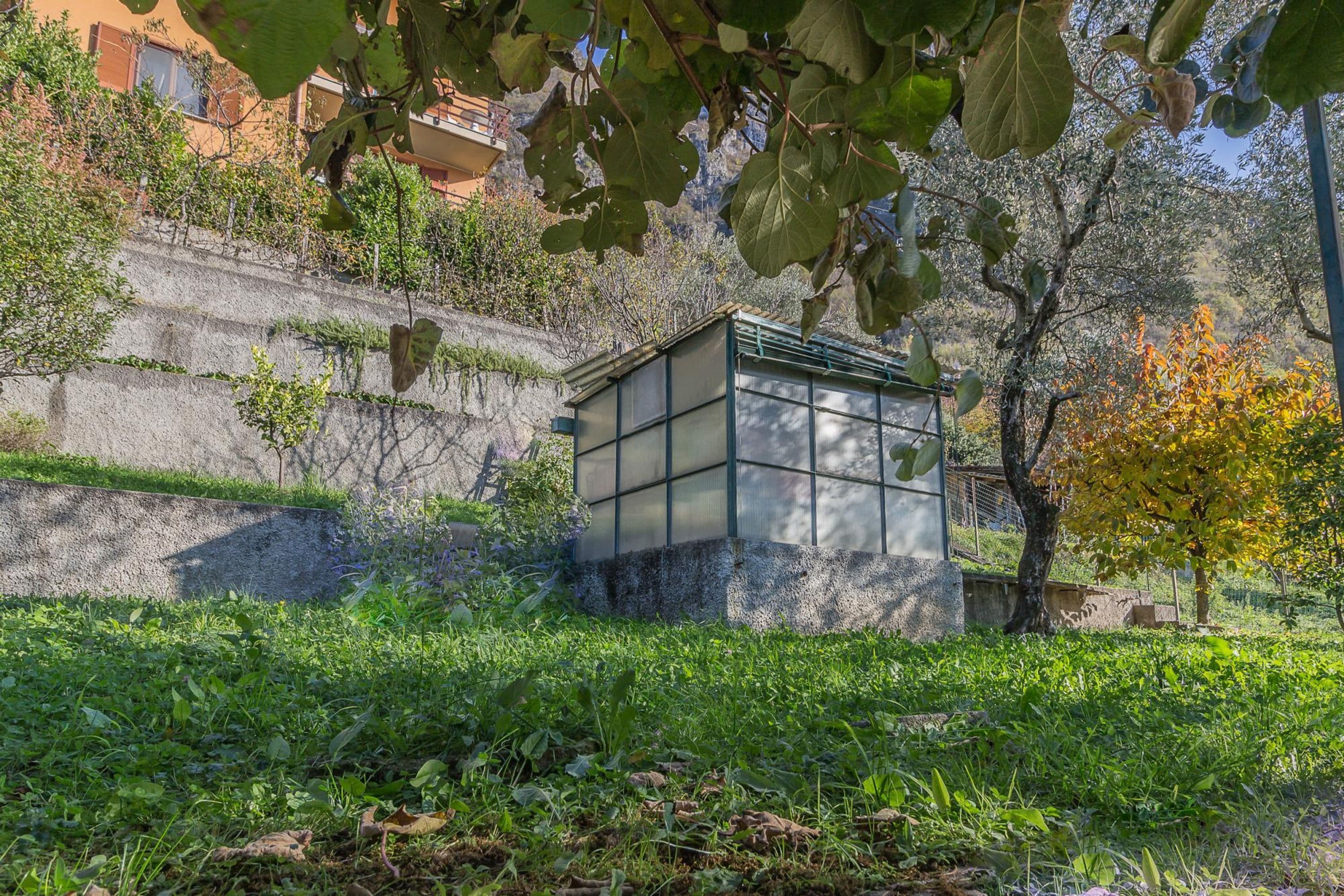 A small, enclosed structure with a glass facade, situated on a grassy slope with a backdrop of trees and a building.