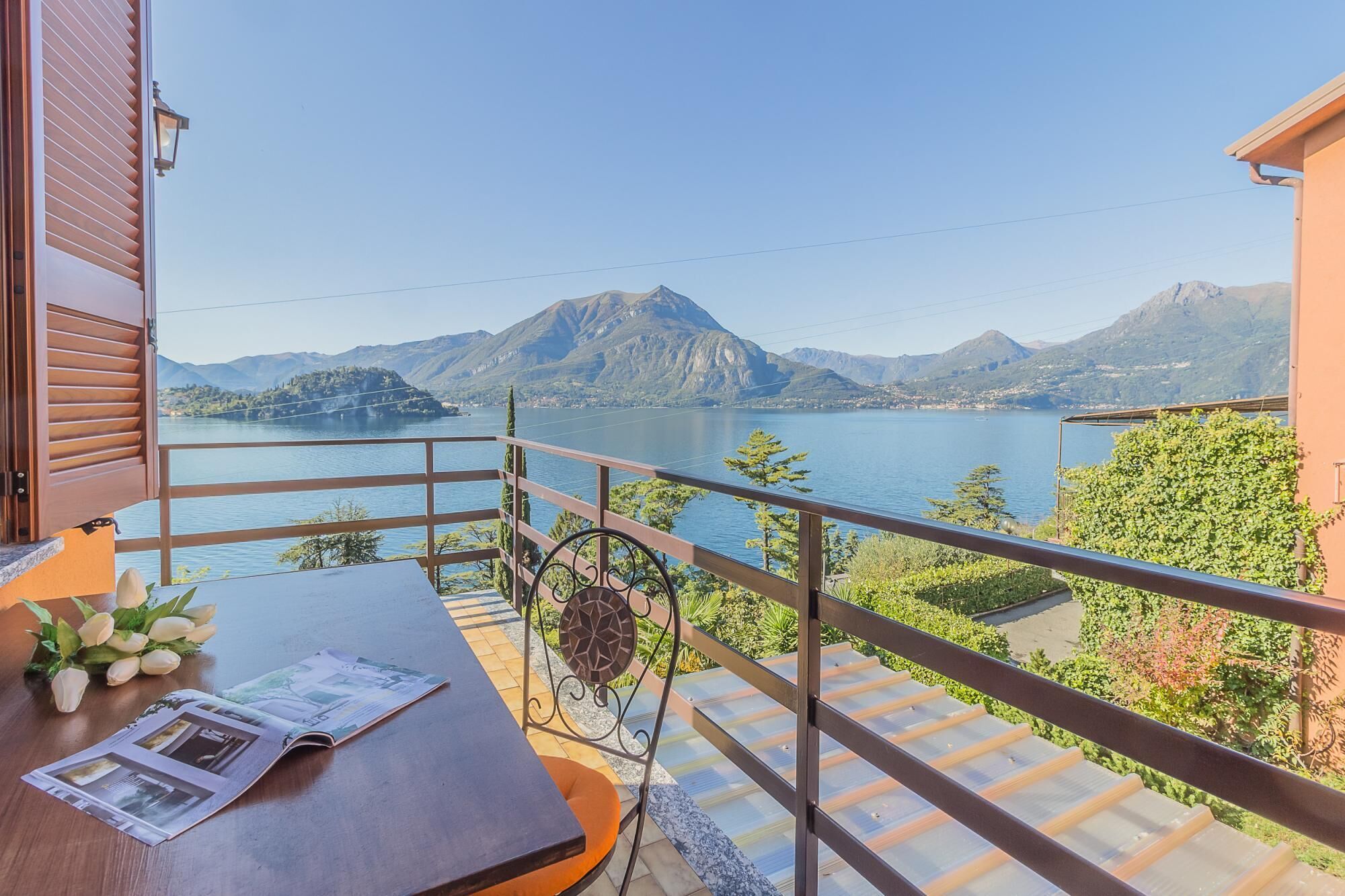 A balcony with a view of a lake and mountains, a table with a newspaper, and a potted plant.