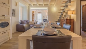 Un coin repas avec une table en bois dressée pour quatre personnes, un canapé et une cuisine équipée d’un four ancien.