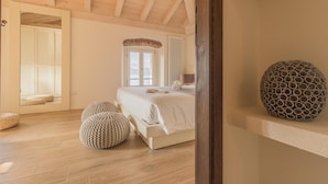 Une chambre avec un plafond en bois, un grand lit, deux poufs en tricot, un objet décoratif rond posé sur une étagère et une fenêtre avec vue.