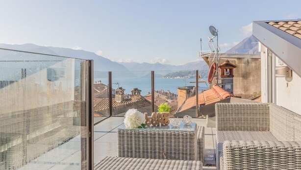 A balcony with wicker furniture, a glass enclosure, and a view of the sea and mountains. - La Terrazza di Marta (Varenna)