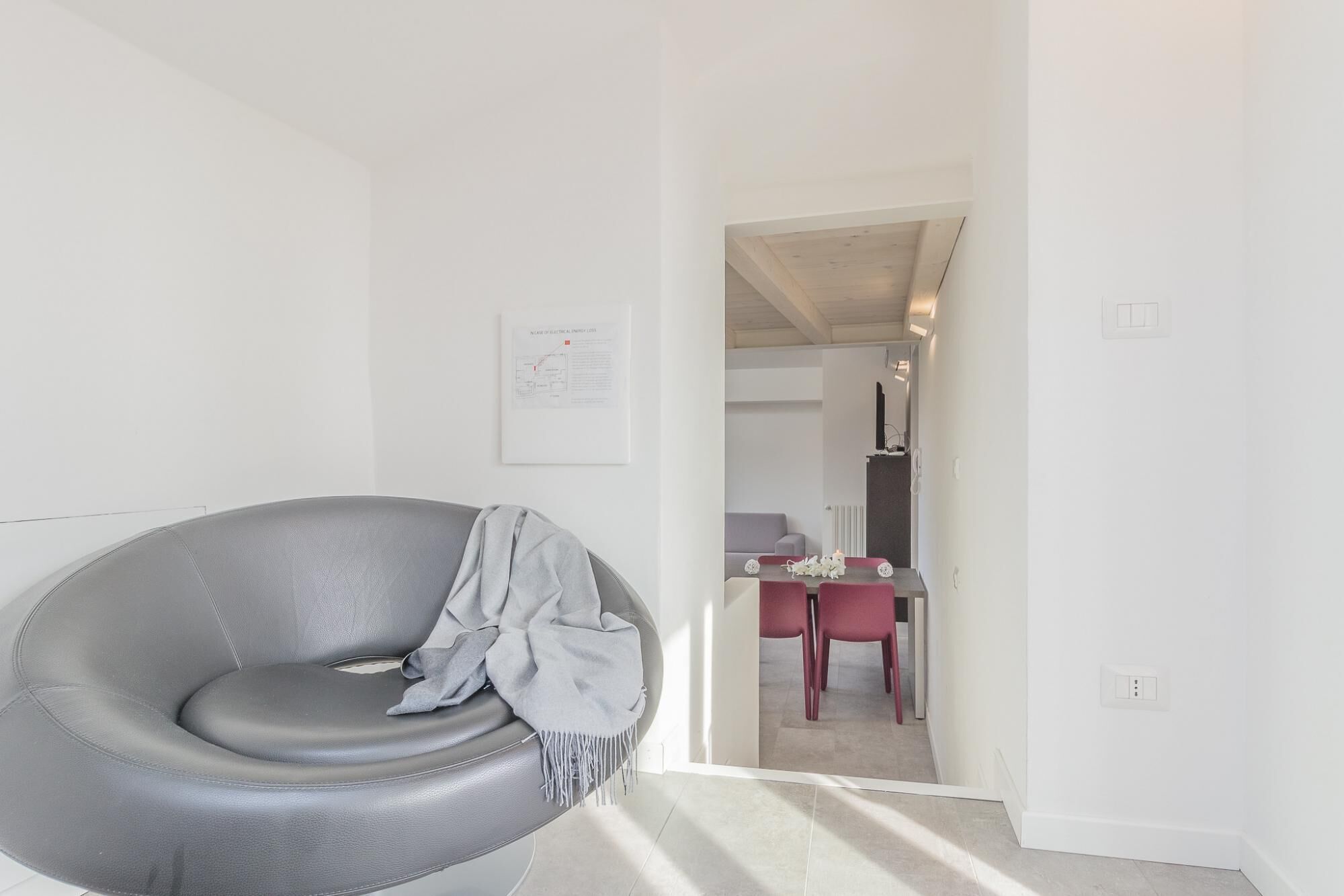 Ein modernes Wohnzimmer mit einem grauen Ledersessel, einem gerahmten Schaltplan an der Wand und Blick in den Essbereich mit einem roten Tisch und Stühlen.