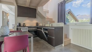 A modern kitchen with a built-in dishwasher, a dining area with a pink chair, and a view of mountains through a window. - La Terrazza di Marta (Varenna)