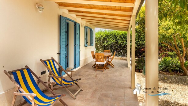A covered outdoor patio with wooden furniture and blue shutters. - Sr-f943-gmar0bct - Country Chic (Noto)