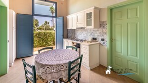 A compact kitchen with a round table and chairs, a window with a view of greenery, and a green door. - Sr-f943-gmar0bct - Country Chic (Noto)