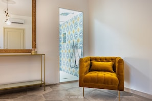 Bathroom features a gold-framed mirror, console table, and tufted chair, with a shower area and patterned glass door.