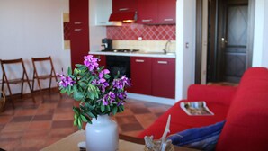 Ein Wohnzimmer mit einem roten Sofa, einem Couchtisch mit einer Vase voller violetter Blumen und einer Küche im Hintergrund mit roten Schränken und einer gefliesten Spritzschutzleiste.