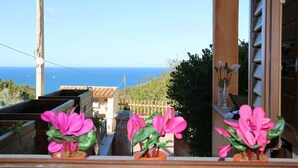 Ein Balkon mit blühenden Kübelpflanzen, Blick auf das Meer und ein Gebäude mit einem Ziegeldach.