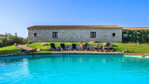 Un edificio de piedra con techo de tejas, piscina y varias sillas de descanso.