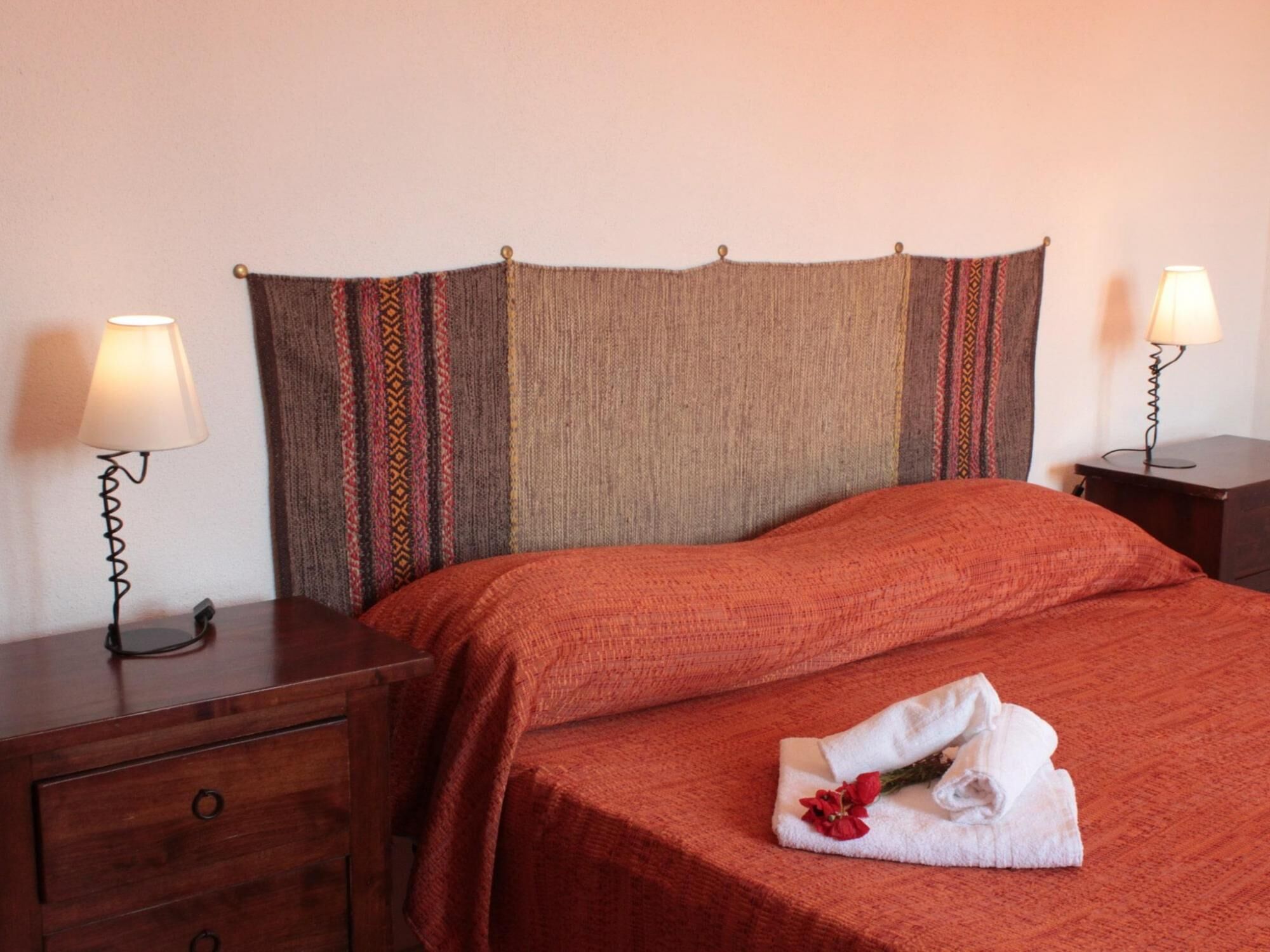 Ein ordentlich bezogenes Bett mit roter Decke, ein Nachttisch aus Holz mit Lampe und ein gefaltetes Handtuch mit Blumen auf dem Bett.