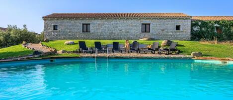A stone building with a tiled roof, a swimming pool, and lounge chairs.
