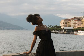 A person in a black dress standing by a waterfront with buildings and a boat in the background. - Il Diamante del Lago (Varenna)
