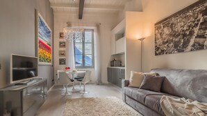 Un salon moderne avec un canapé gris, un téléviseur, un coin repas comprenant une table et des chaises blanches, et une grande fenêtre offrant une vue sur une montagne.