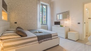 Une chambre à coucher avec un grand lit, une commode, un miroir et une fenêtre donnant sur des immeubles.