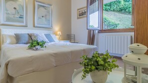 A bedroom with a bed, a bedside table with a lamp, a window with a view of greenery, and framed artwork on the wall. - Villa Lisander Varenna (Varenna)