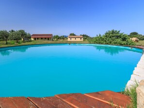 A clear blue swimming pool surrounded by a stone deck, with a house and greenery in the background. - d Pietraia 11B Trilo (Badesi)