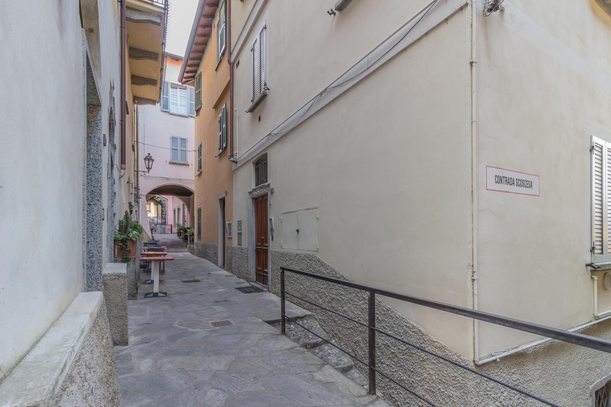 A narrow alleyway with stone pavement, a metal railing, and buildings with doors and windows.