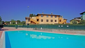 A swimming pool with a clear blue water surface, surrounded by a green fence. In the background, there is a yellow building with multiple windows and balconies. - Appartamento Tammaro (Sirmione)