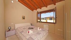 A bedroom with a wooden ceiling, a bed with a quilt, a bedside table with a lamp, a window with a view of buildings, and a radiator. - Appartamento Tammaro (Sirmione)