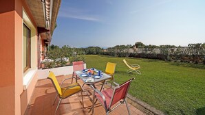 An outdoor patio with a table set for two, surrounded by chairs, overlooking a well-maintained lawn and a distant view of buildings.