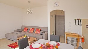 Een moderne woonkamer met een grijze bank, een eettafel gedekt met borden, glazen en fruit. Op de muur hangt een klok en er staat een houten kast met keukengerei.
