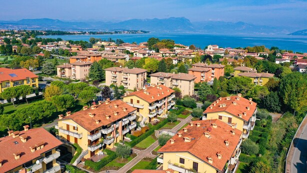 A residential area with multi-story buildings, some with balconies, and a view of a lake and mountains in the distance. - Villaggio dei Fiori - Viola (Sirmione)
