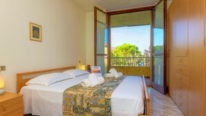 A double bed with a patterned bedspread, a bedside table with a lamp, and a window with a view of trees and buildings. - Villaggio dei Fiori - Viola (Sirmione)