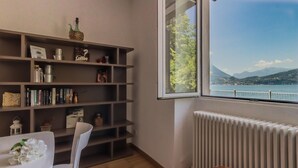 A room with a bookshelf, a table with a white chair, and a view of a lake and mountains through a window. - Varenna Beach Perledo (Perledo)
