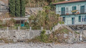 A stone wall with a metal gate, a building with green shutters, and a river with ducks. - Varenna Beach Perledo (Perledo)