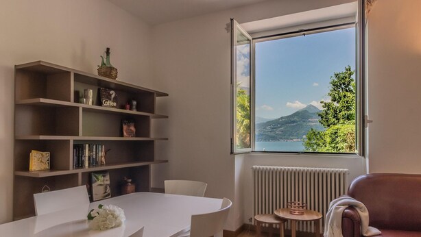A room with a white table, white chairs, a brown sofa, a bookshelf, and a window with a view of a mountain and trees. - Varenna Beach Perledo (Perledo)