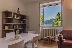 A room with a white table, white chairs, a brown sofa, a bookshelf, and a window with a view of a mountain and trees. - Varenna Beach Perledo (Perledo)