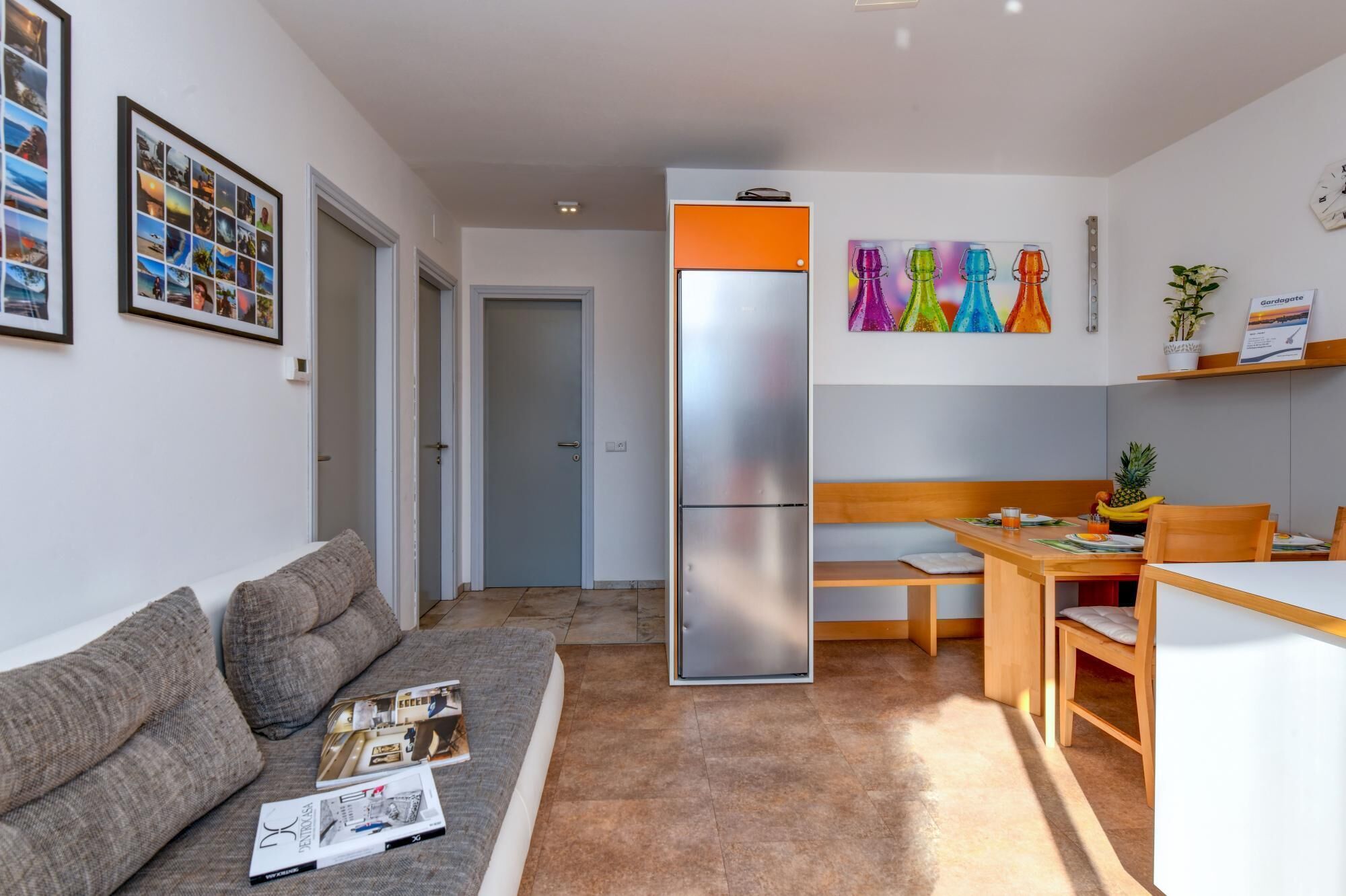 A modern living room with a grey sofa, a dining table with chairs, a kitchen area with a fridge, and a hallway leading to a door.