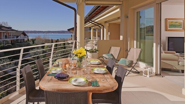 Une table à manger installée sur un balcon avec vue sur la mer, entourée de chaises en osier et protégée par une rambarde en verre.