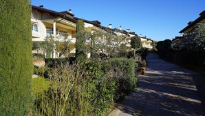 Une rangée d’immeubles à plusieurs étages, dotés de balcons, entourée d’un espace vert bien entretenu et bordée d’un chemin pavé.