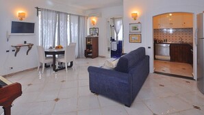 A living room with a blue sofa, a dining area with a table and chairs, a TV mounted on the wall, and a kitchen with cabinets and a refrigerator. - Villa With Terrace and sea View in Ravello ID 3196 (Ravello)