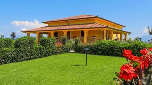 A yellow villa with a tiled roof, surrounded by well-maintained hedges and a grassy lawn.