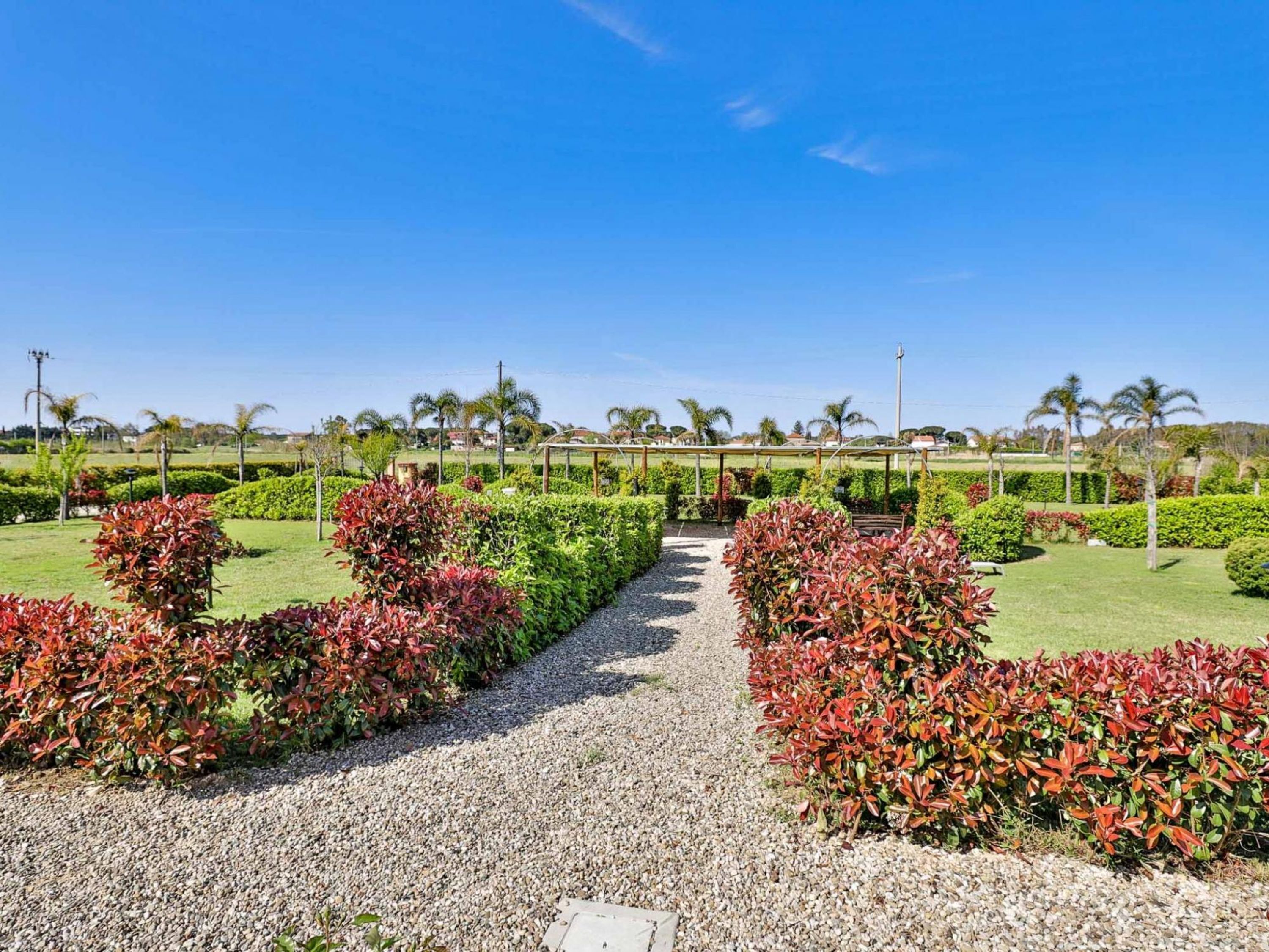 A pathway leading through a well-maintained garden with neatly trimmed hedges and palm trees in the background.