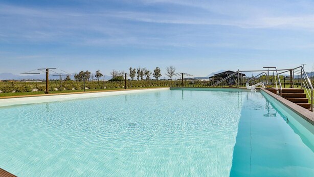 A clear blue swimming pool with a wooden deck in the foreground, surrounded by a landscaped garden and a distant mountain range. - I Giardini Dell Olimpo - Zeus (Capaccio-Paestum)