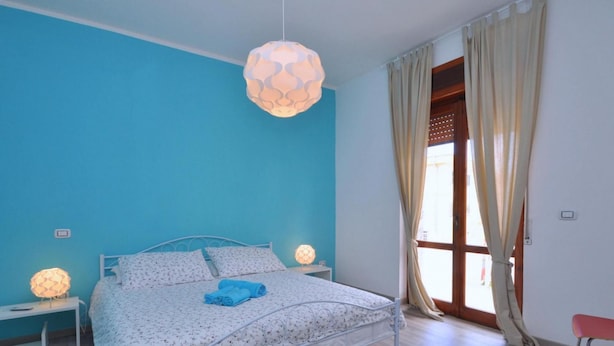 Ein Schlafzimmer mit einem Bett, zwei Nachttischlampen, einer Pendelleuchte, einem Fenster mit Vorhängen und einem roten Sessel.