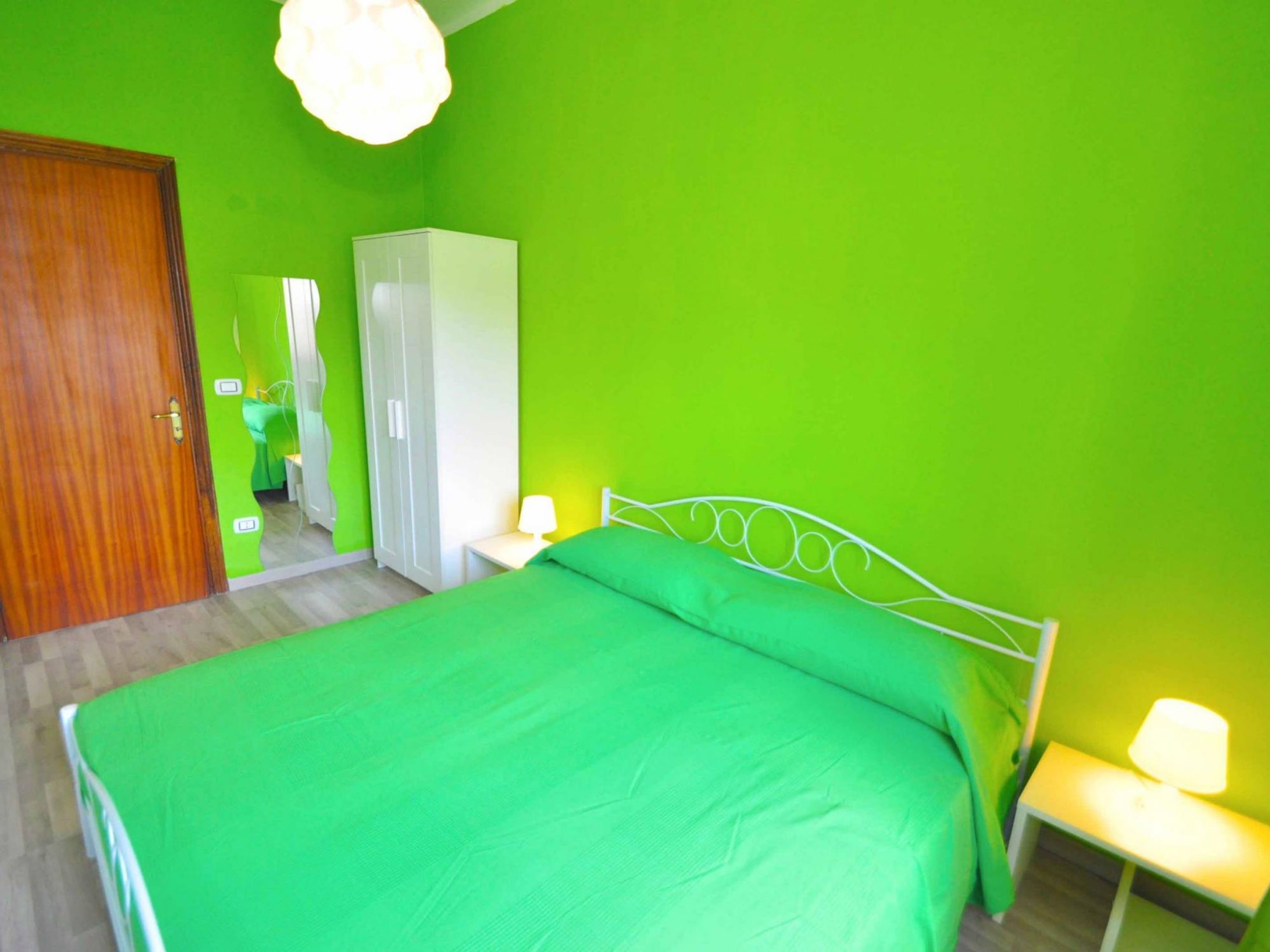 A room with a bed, a wardrobe, a mirror, and a nightstand with a lamp.