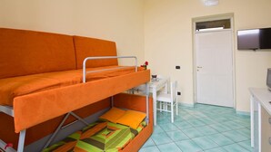 Quarto com beliche, uma mesa pequena, uma cadeira, televisão e porta.