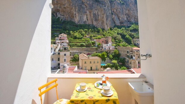Un balcon avec une table dressée pour deux personnes, offrant une vue sur un village aux maisons à étages et sur une falaise imposante.