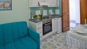 Une cuisine compacte avec des meubles blancs, une petite cuisinière et un évier. On y trouve un canapé de couleur turquoise et une table à manger sur laquelle sont posés quelques objets.